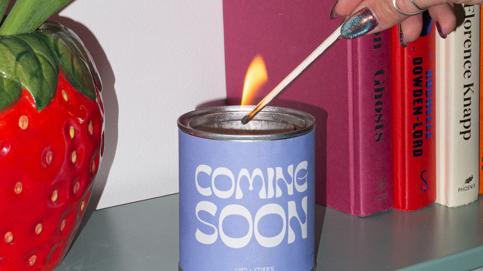Hand lighting a candle labeled 'Coming Soon' on a shelf with books and a strawberry-shaped vase.